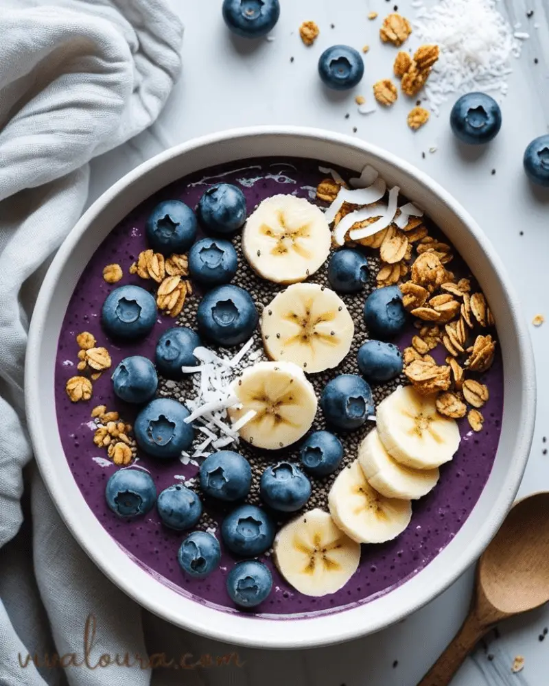 Blueberry smoothie bowl topped with fresh blueberries, banana slices, chia seeds, coconut, and granola on a white surface